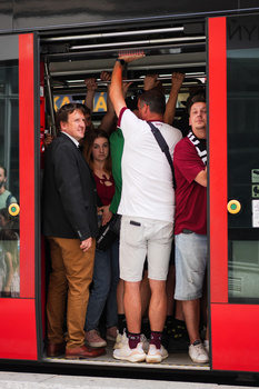 Crowded public transit vehicle with passengers standing near open doors framed by bright red panels.