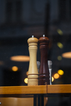 Light and dark wooden salt and pepper mills standing upright on a wooden surface with blurred warm bokeh lights in the background.