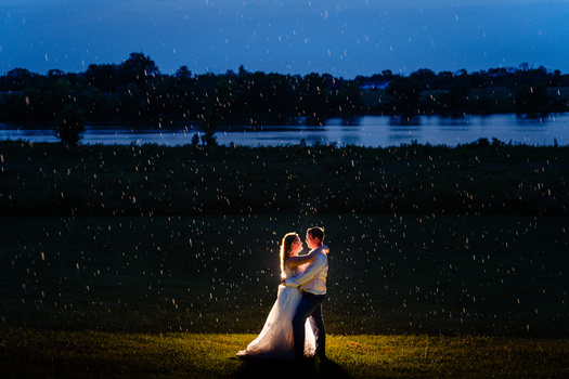 Bride and groom embracing in heavy rain at dusk with a glowing light source illuminating them against a dark landscape and water backdrop.