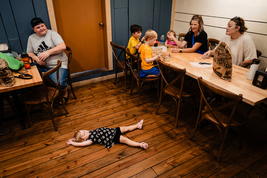 Child lying on wooden floor during a group gathering in an indoor space with multiple people seated at tables.