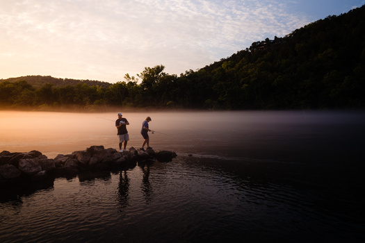 Two people standing on rocks at the edge of a misty lake at sunrise, surrounded by forested hills.