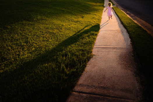 Child in pink dress walking on concrete sidewalk beside grass field during golden hour.