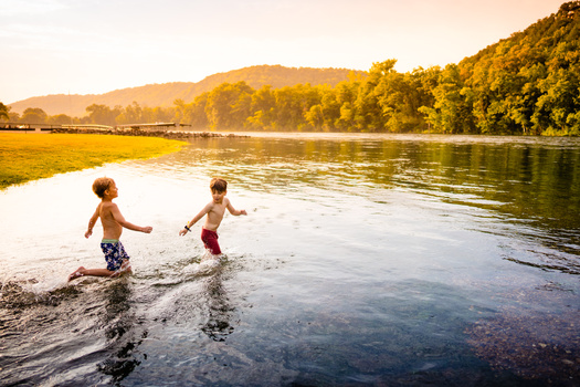 Two shirtless children wading into a river at golden hour with forested hills in the background.