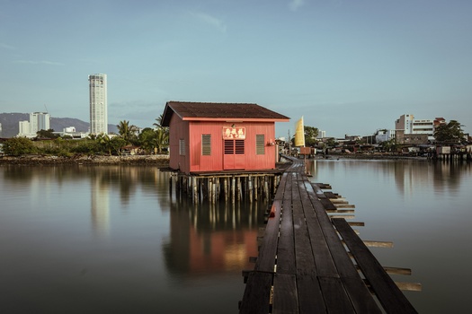 Red wooden structure on wooden pilings extending into calm water with city skyline in background.