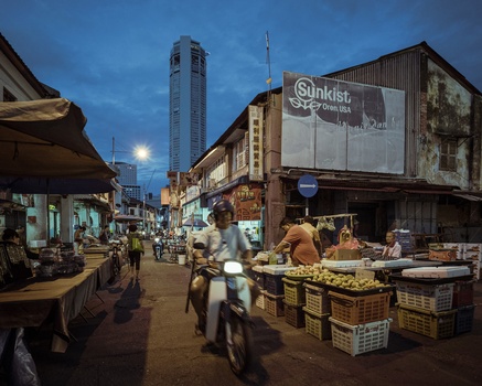 Evening street market scene with vendor stalls, pedestrians, and illuminated high-rise building in background.