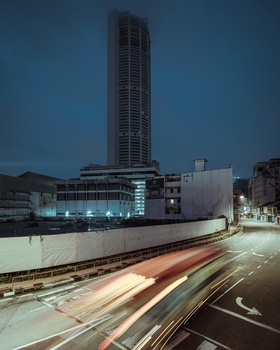 Tall residential tower illuminated at dusk with light trails from vehicle traffic in foreground.