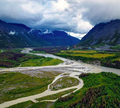 Braided river valley with winding waterways, forested slopes, and distant snow-capped mountains under dramatic cloudy sky.