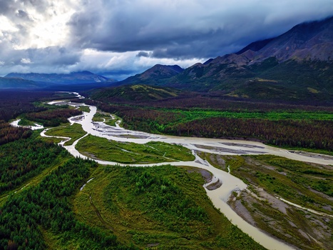 Aerial view of a winding braided river cutting through forested valleys beneath dramatic storm clouds and mountains.