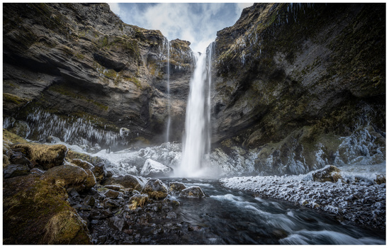 Waterfall cascading between dark rocky cliffs into a stream with ice formations below.