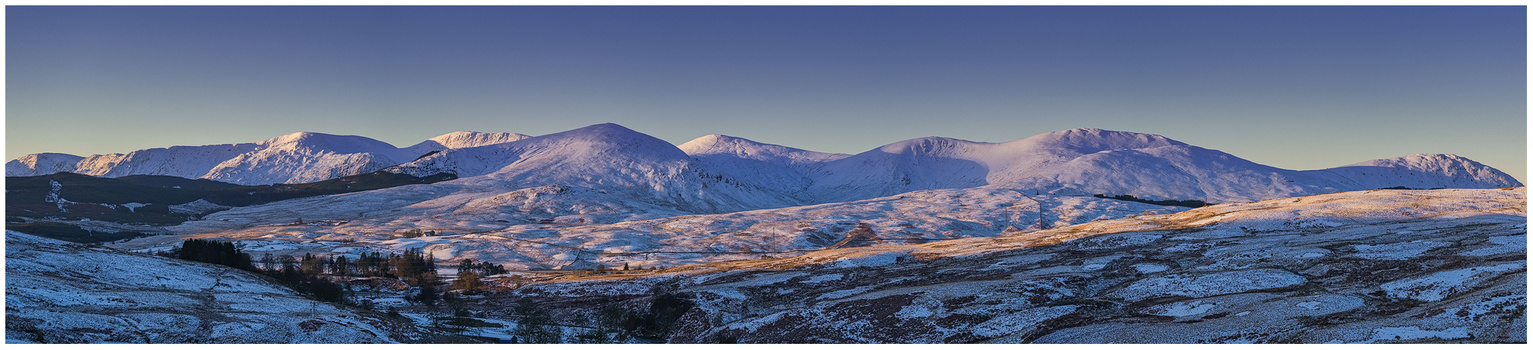 Snow-covered mountain range with golden sunlit peaks at dawn or dusk under clear blue sky.