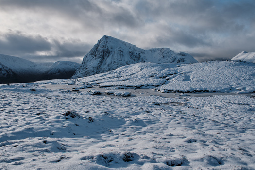 Snow-covered mountain landscape with dramatic cloudy sky and rocky terrain in foreground.