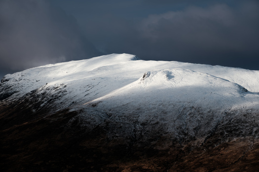 Snow-covered mountain peak with dramatic dark storm clouds overhead and sunlit slopes.