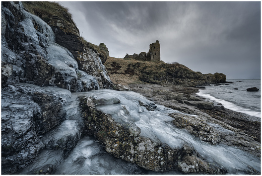 Frozen coastal rocks with icy formations in the foreground, rocky shoreline and ruined stone structure in the distance under overcast sky.