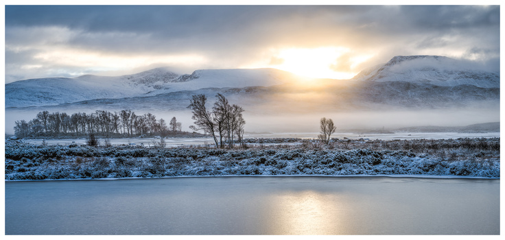 Frozen lake with snow-covered mountains and bare trees at sunrise, golden light breaking through clouds.