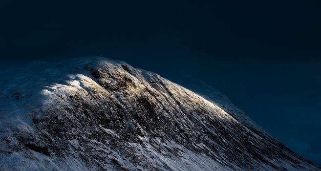Snow-covered mountain peak against deep blue sky with dramatic side lighting.