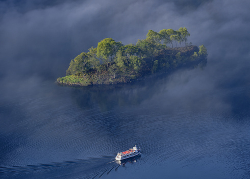 Small cargo ship navigating calm waters near a densely forested island beneath overcast skies.
