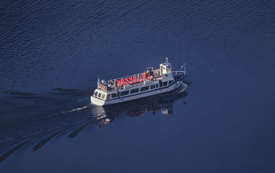 Aerial view of a passenger ferry with red and white hull cutting through deep blue water, leaving a distinctive wake pattern behind.