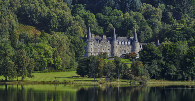 Medieval castle with multiple turrets reflected in calm river, surrounded by dense green forest.