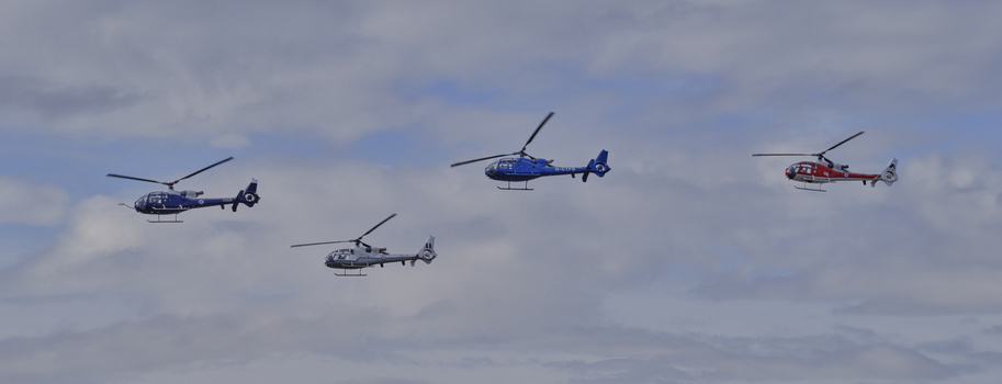 Four helicopters in formation flying against a cloudy sky, three with dark fuselages and one blue, one red.