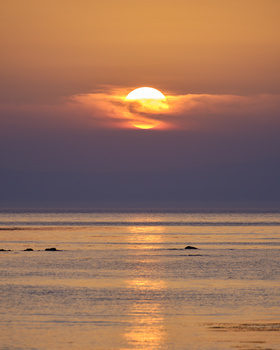 Large sun setting over a calm ocean horizon beneath a gradient sky of orange, pink, and purple tones.