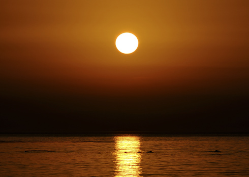 Bright sun reflecting on calm ocean water at sunset, with golden light creating a path across the horizon.
