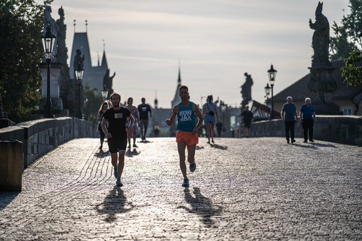 Runners jogging across a historic cobblestone bridge with Gothic architecture and statues lining both sides.