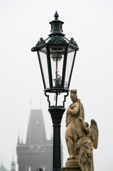 Ornate black street lamp with golden angel statue in foreground, cathedral spire visible through fog in background.