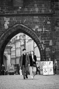 Two people walking through a stone archway toward a city street, captured in black and white.