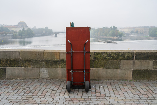 Red industrial cart positioned against a stone wall overlooking a river and distant cityscape.