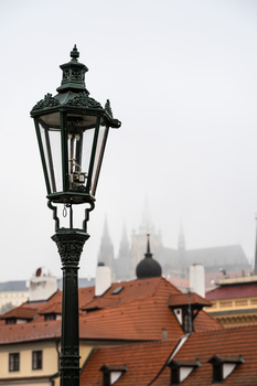 Ornate black street lamp in foreground with European architecture and spires blurred in misty background.