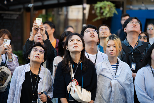 Group of people looking upward during daytime outdoor event, some holding phones and cameras.