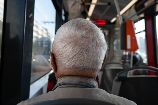 Rear view of an elderly man with white hair sitting on public transportation, looking toward the front of the bus.