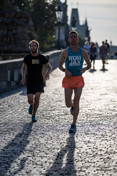 Two runners jogging on a cobblestone bridge with historic architecture and crowds visible in the background.