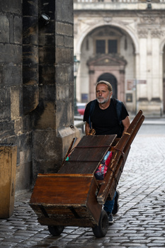 Man in glasses pushing a wooden handcart through a European cobblestone street with classical architecture in the background.