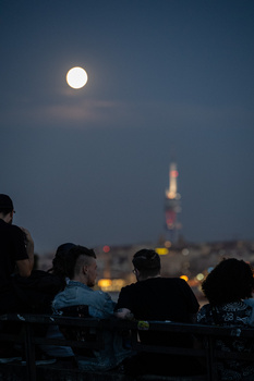 Full moon rising above a distant illuminated tower while silhouetted figures watch from the foreground.
