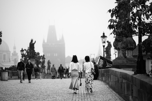 Two women walking away on a cobblestone bridge toward Gothic architecture shrouded in fog.