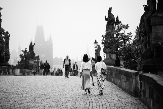Two women walking away on a historic stone bridge lined with statues, with a Gothic cathedral silhouetted in misty background.