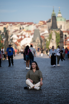 Woman sitting cross-legged on a cobblestone plaza with a historic European city and church spires in the background.