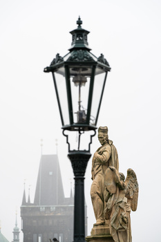Golden angel statue beside a vintage street lamp with Gothic cathedral spires visible in misty background.