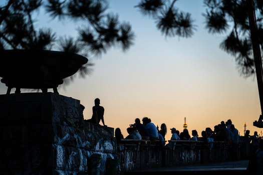Silhouetted figures gathered on a rocky overlook at sunset, with pine trees framing the scene.