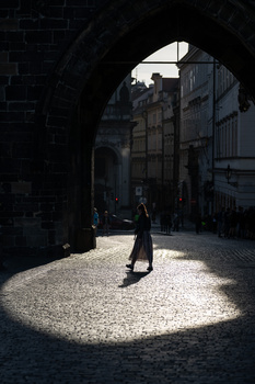 Figure walking through a sunlit archway in a narrow cobblestone passage between historic stone buildings.
