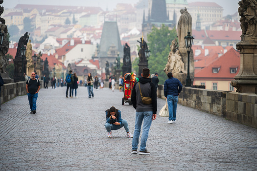 Photographer crouching to capture a street scene on a historic European bridge with pedestrians and cityscape.