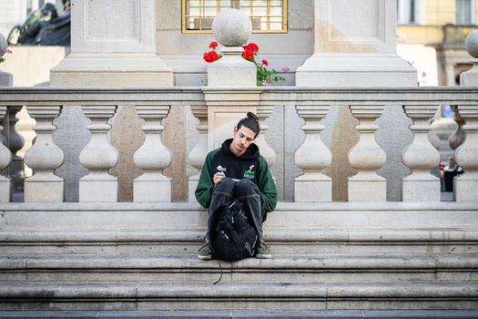 Person sitting on wide stone steps wearing a green jacket and black backpack, positioned below ornamental balustrade.