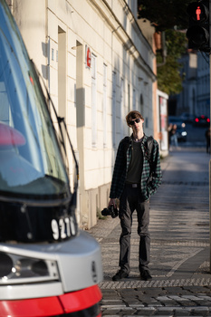 Person in dark clothing standing on an urban street next to a parked tram, with classical buildings and storefronts visible behind.