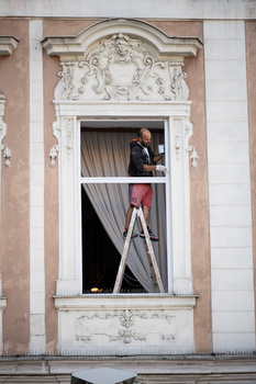 Ornate window with decorative stone surround on classical building facade, showing workers inside with ladder and equipment.