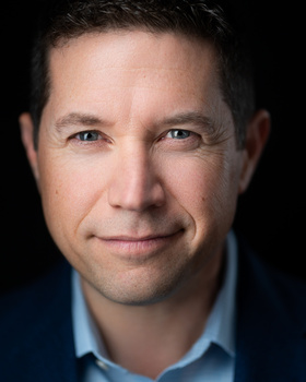 Professional headshot of a man with short dark hair wearing a light blue button-up shirt against a black background.