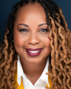 Professional headshot of a woman with blonde ombré locs against a dark blue background.