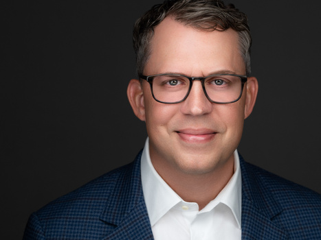 Professional headshot of a man wearing glasses and a blue plaid blazer over a white shirt.