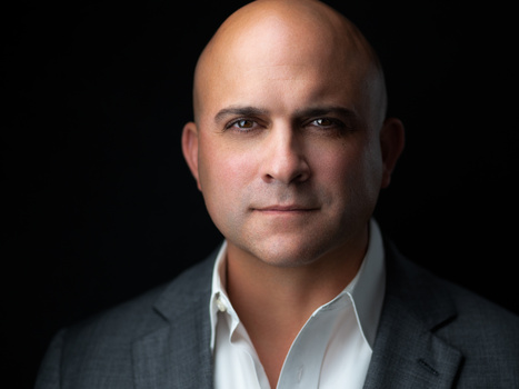 Professional headshot of a bald man in a dark blazer and white shirt against a black background.