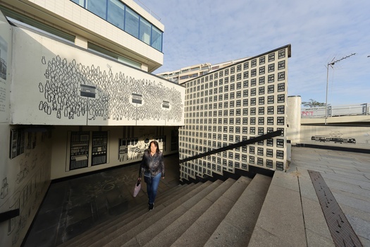 Woman walking down concrete steps toward modernist buildings with geometric facades under daylight.